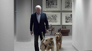 A dapper man in a suit walks down an elegant hallway with his dogs beside him