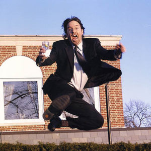 Gerald Jones, dressed in a suit and holding a drink, jumping into the air
